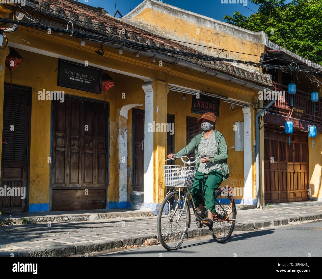 Donna che indossa un cappello da sole e una maschera in bicicletta su una strada storica della città Foto Stock