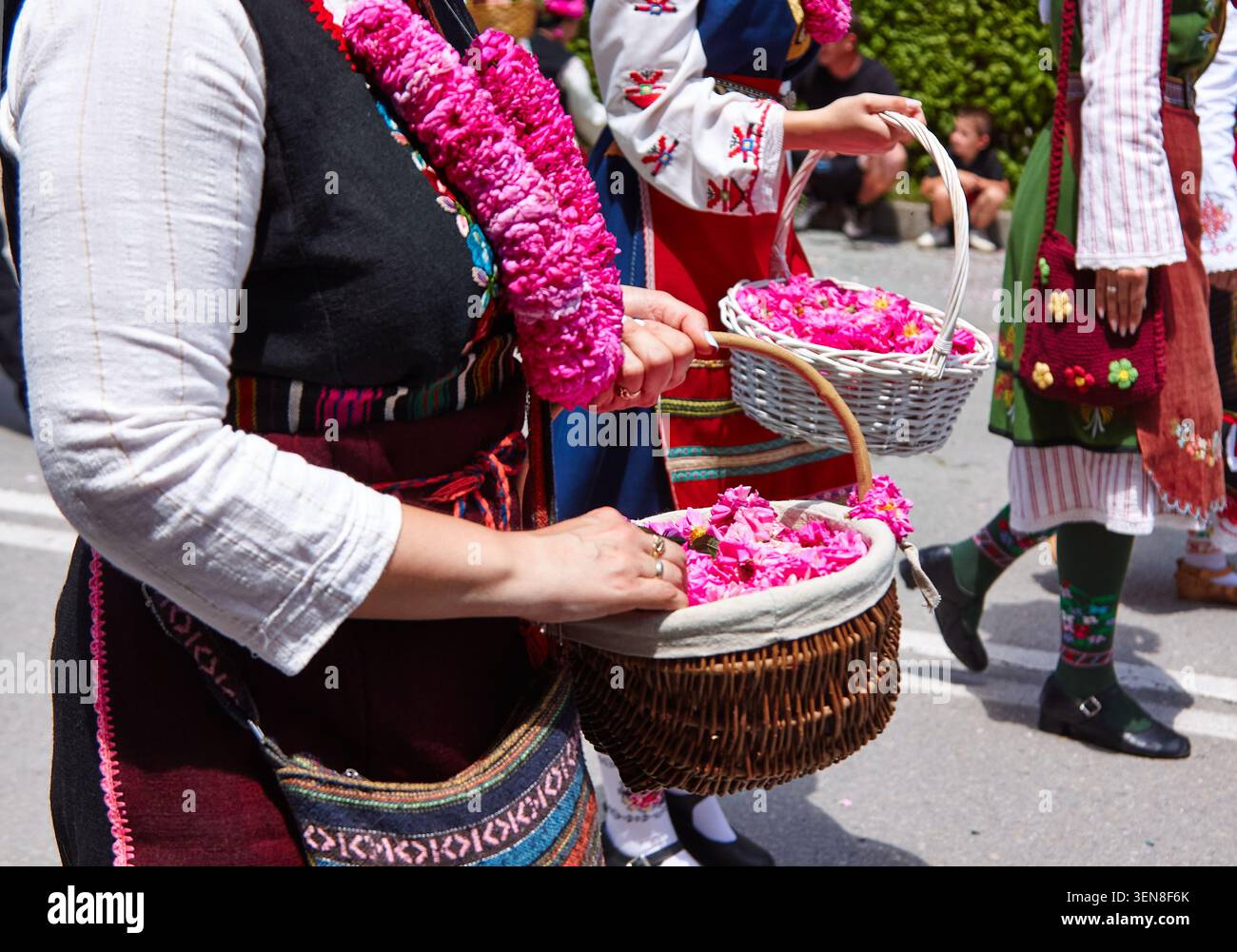 Una donna vestita con abiti tradizionali tiene in mano cesti pieni di petali di fiori di rosa rosa durante una sfilata culturale in una strada vivace Foto Stock