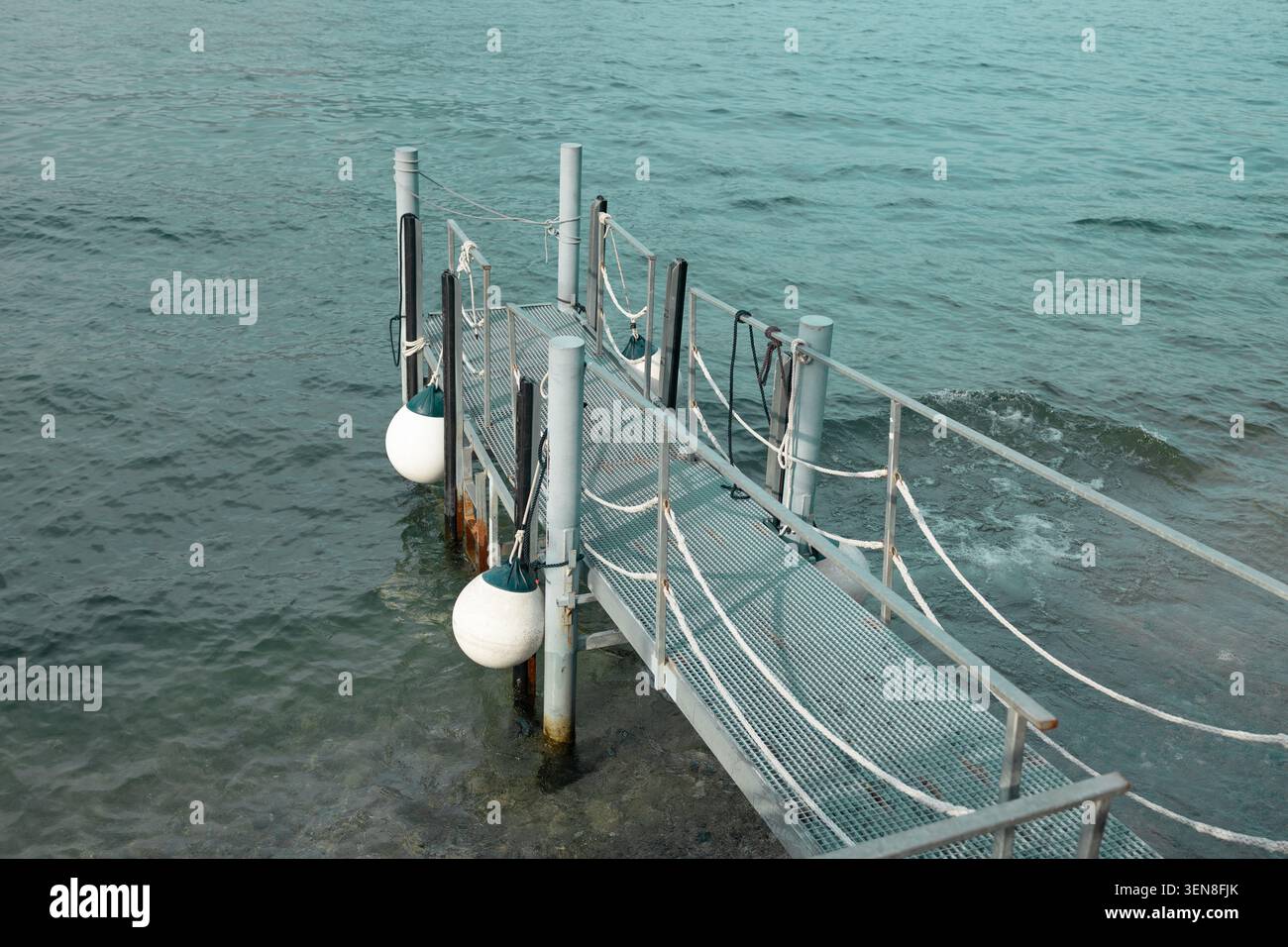 Vista ad angolo alto di un piccolo molo galleggiante o di una passerella con parafanghi bianchi rotondi su un lago turchese calmo in una giornata di sole. Foto Stock