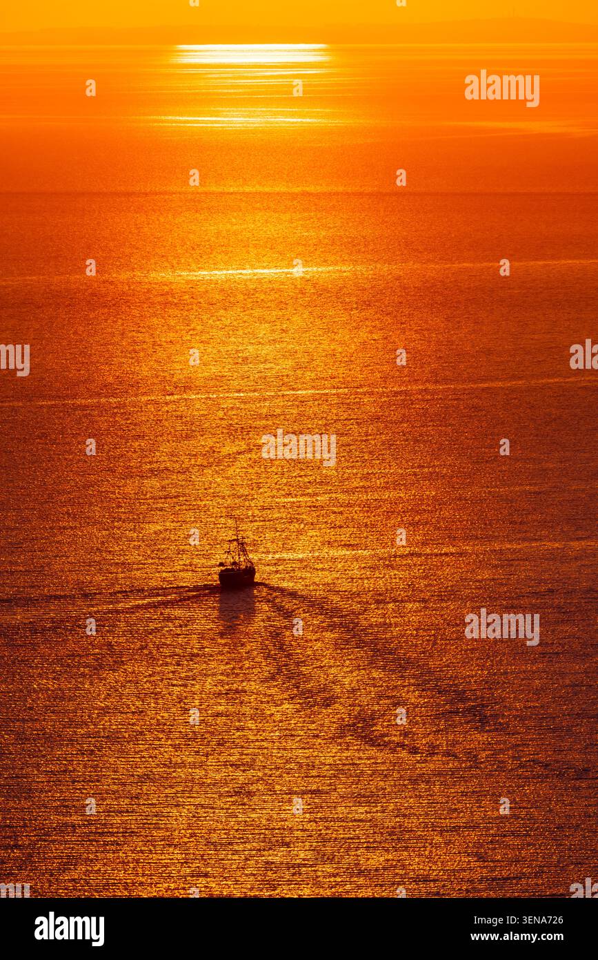 Piccola nave che attraversa il mare dorato al tramonto con un lungo riflesso e la luce arancione del sole Foto Stock