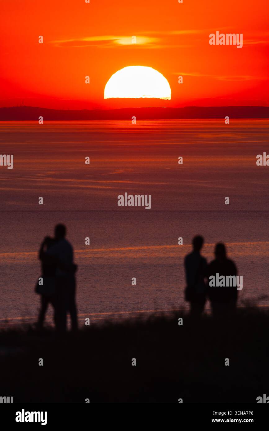 Sagome che guardano il tramonto sul mare calmo con la barca all'orizzonte luminoso Foto Stock
