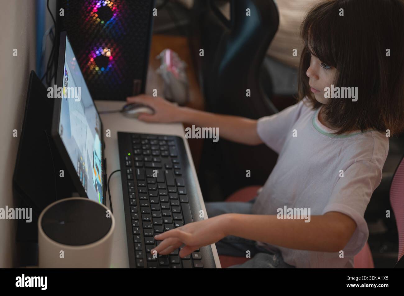 Un bambino siede su una scrivania bianca con un computer, concentrato sul gioco. La camera è dotata di attrezzatura da gioco e di un'atmosfera rilassata. Foto Stock