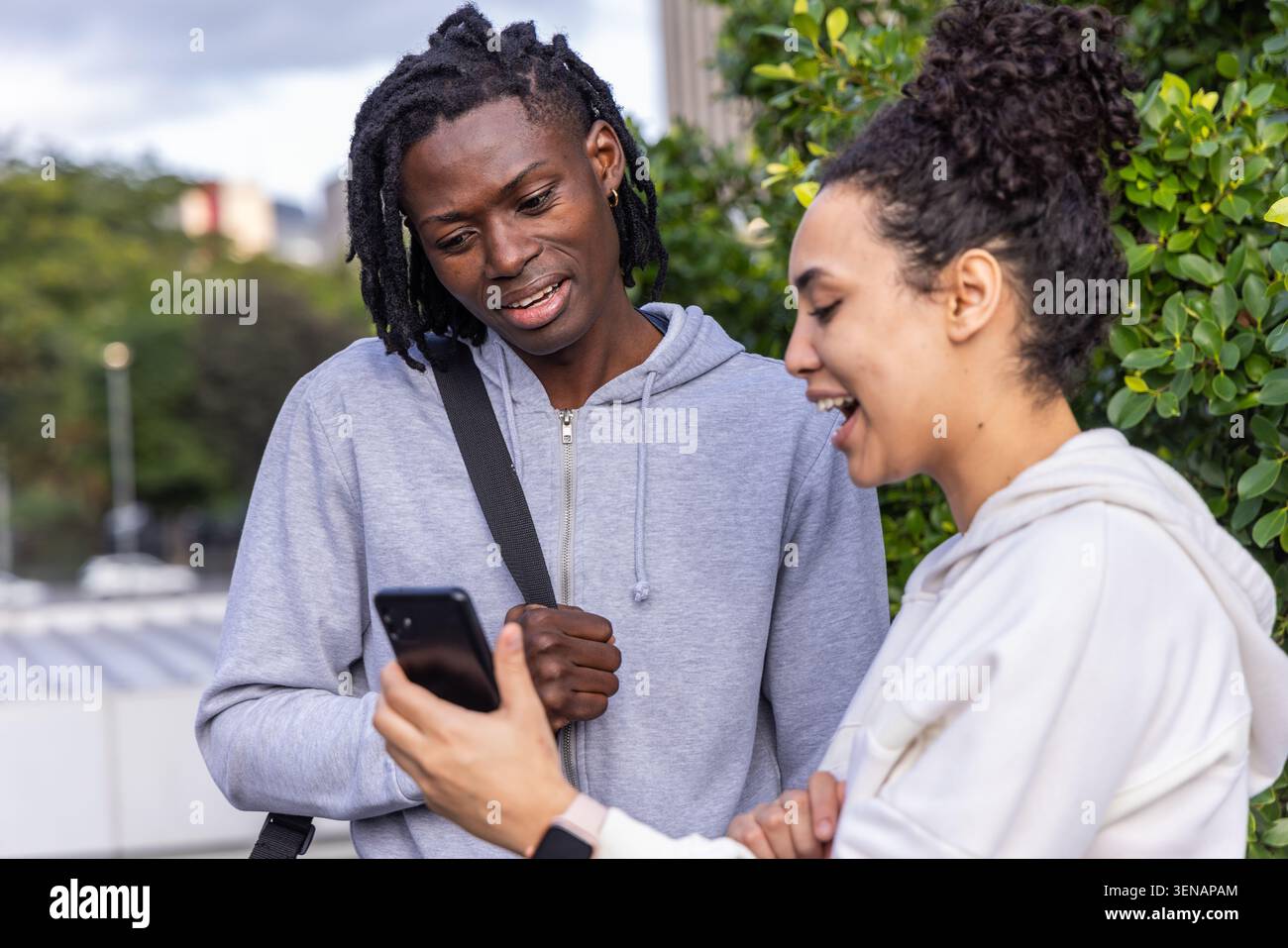 Due amiche in piedi accanto alla ringhiera sul tetto, che condividono il telefono, indossano felpe con cappuccio, orologio, cinturino Foto Stock