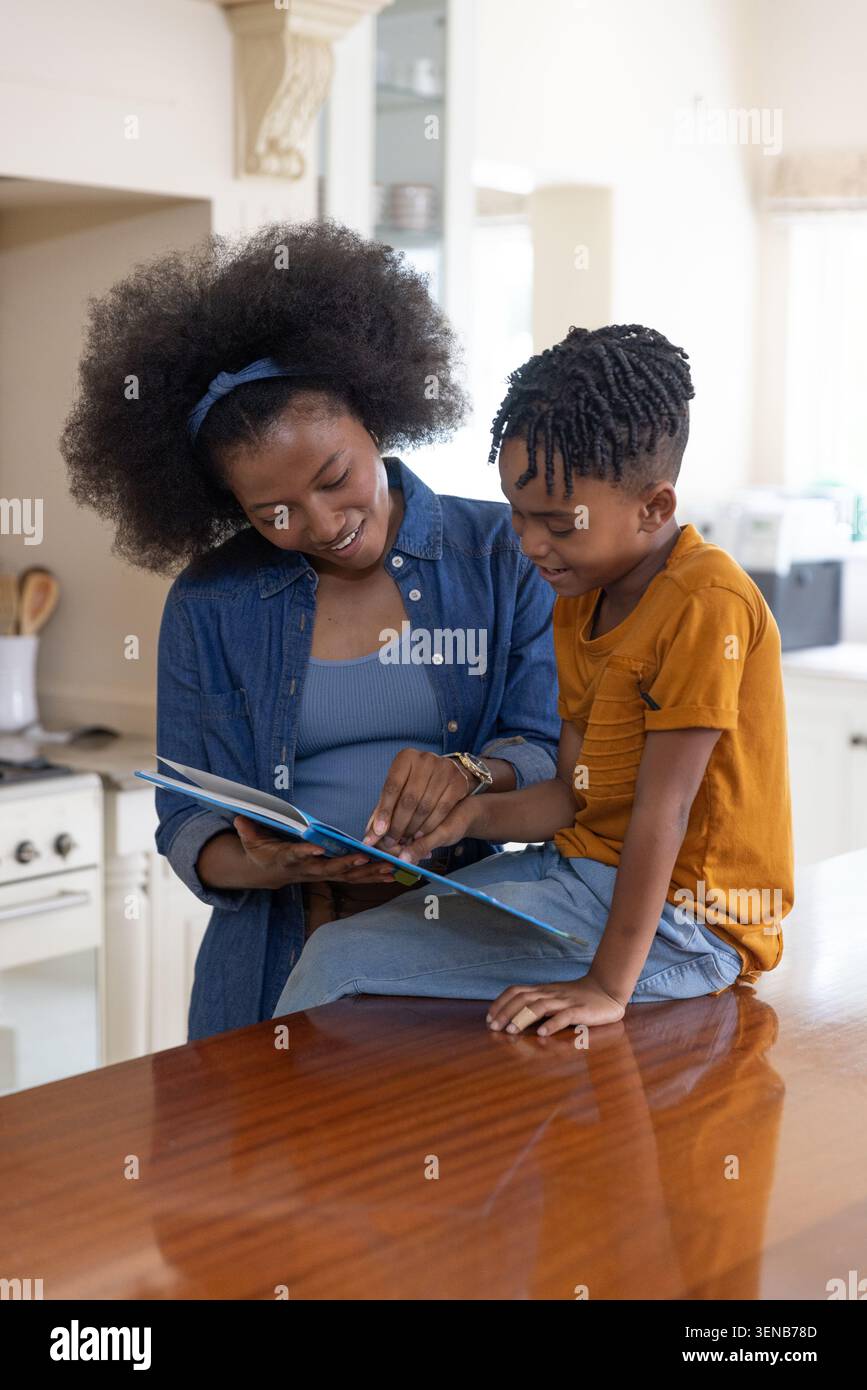 Madre e figlio afroamericani seduti al banco della cucina in legno lucido leggendo un libro illustrato Foto Stock
