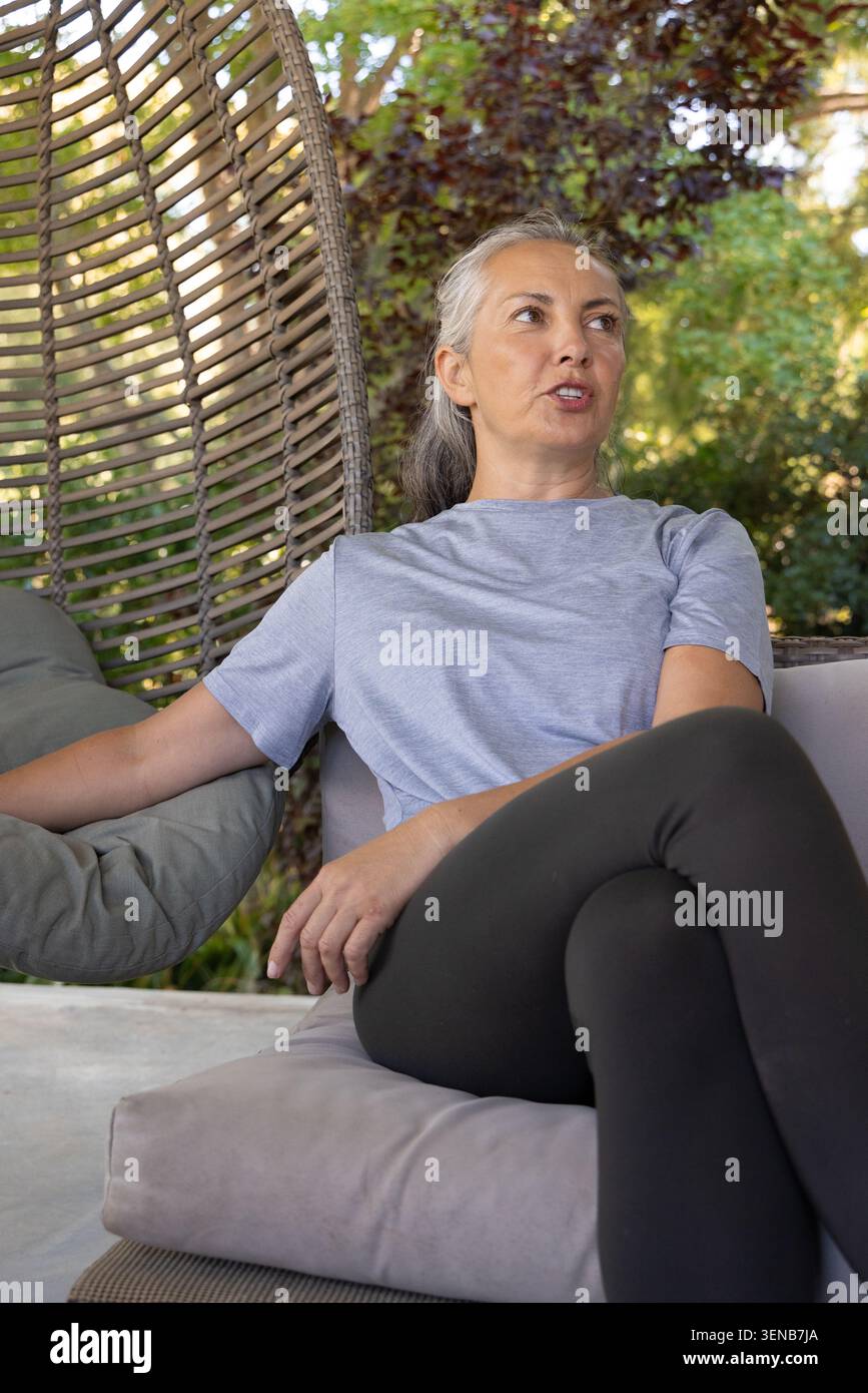 Donna anziana seduta su una sedia a uovo intrecciata sul patio con cuscino, indossa una maglietta, leggings neri, parla Foto Stock