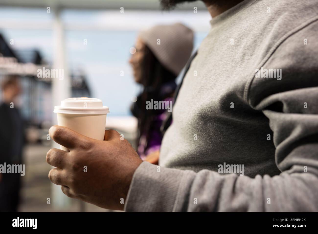 Primo piano del passeggero dell'aeroporto che tiene in mano una tazza di caffè da asporto accanto ai viaggiatori che camminano su sfondo sfocato. Contenitore per bevande in mano turistica che naviga nel centro di trasporto dell'aeroporto Foto Stock