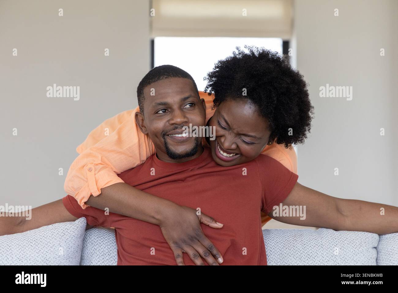 Coppia afroamericana che ride mentre si appoggia su un divano di colore chiaro a casa con tendina parasole Foto Stock