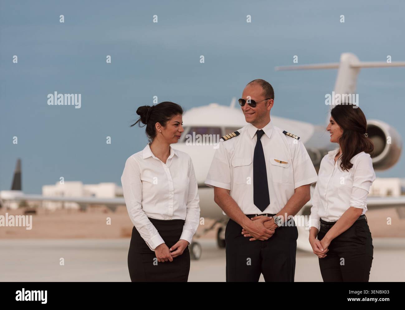 Cordiale equipaggio privato con pilota sorridente e due assistenti di volo in uniforme bianca che parlano sull'asfalto dell'aeroporto al crepuscolo dorato accanto a un'azienda Foto Stock