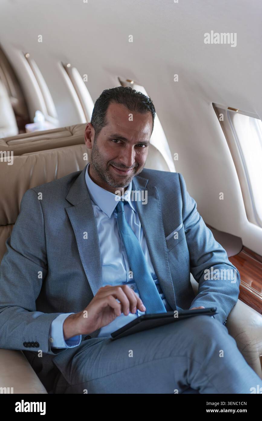 Uomo d'affari del Medio Oriente sorridente in tuta grigia che utilizza un tablet digitale a bordo di una lussuosa cabina jet privata con luce soffusa sul finestrino, ideale per i dirigenti Foto Stock