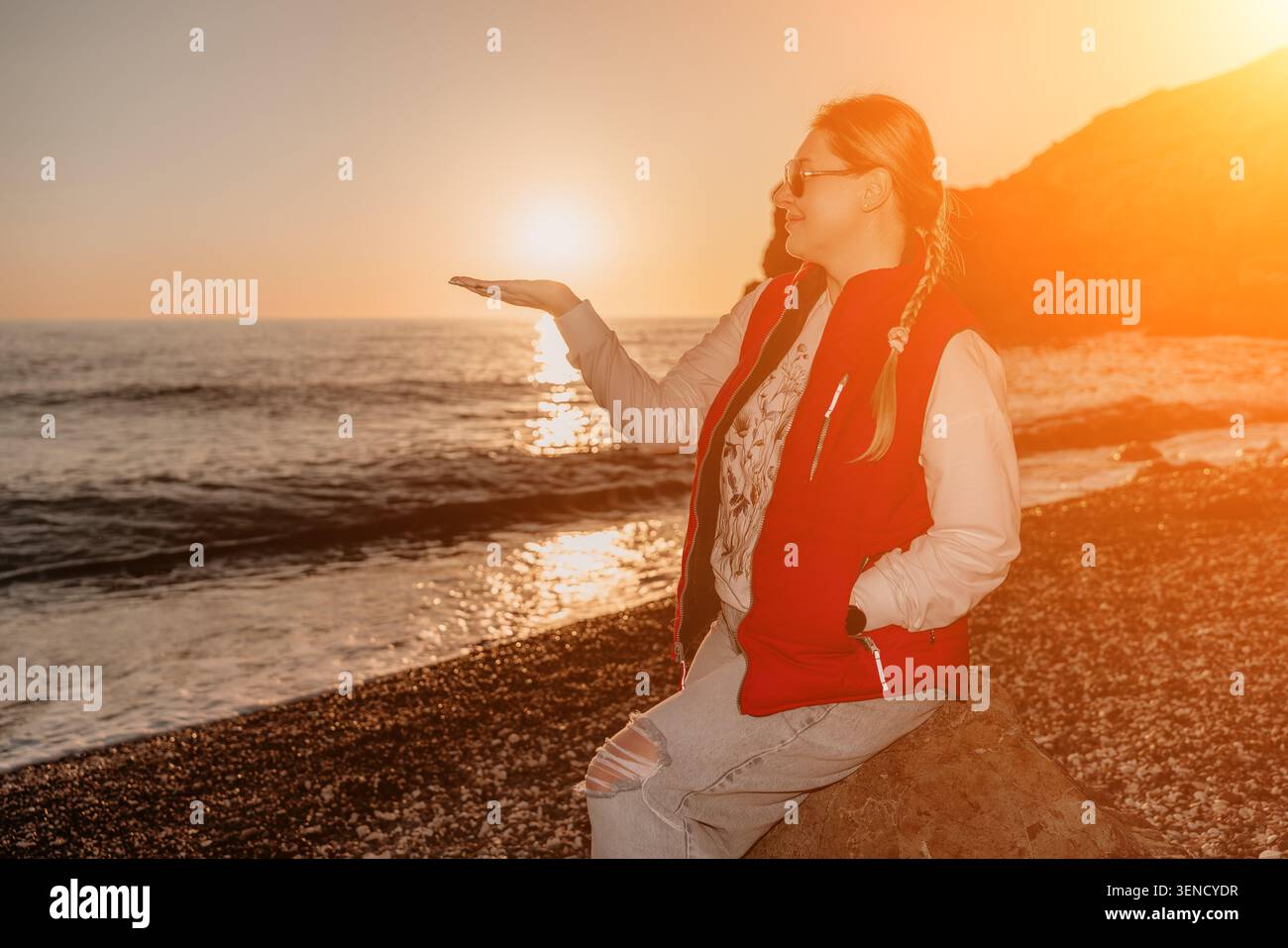 Spiaggia al tramonto donna che cattura una prospettiva giocosa tenendo il sole in mano mentre è seduta su una spiaggia rocciosa Foto Stock