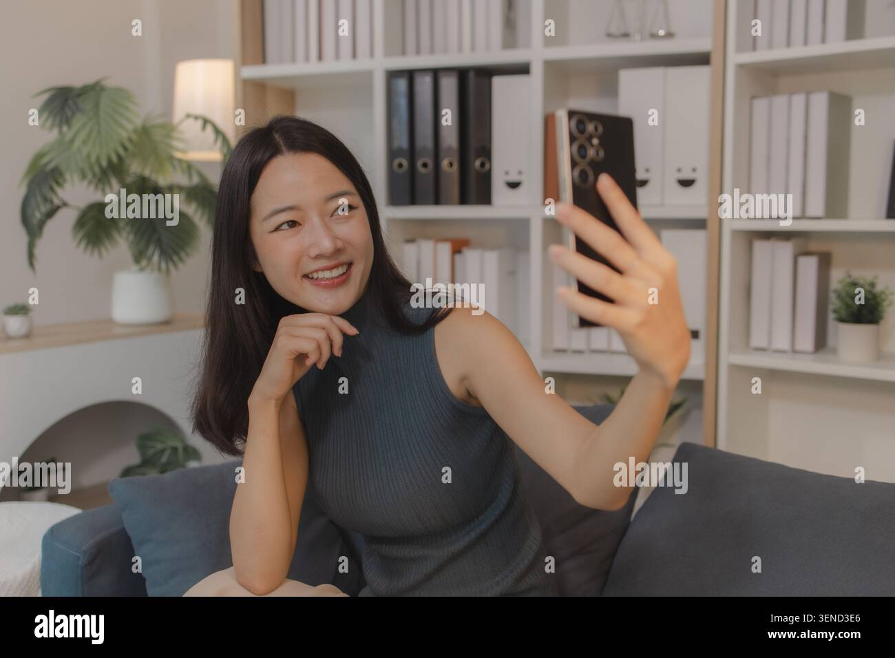 Giovane donna asiatica seduta sul divano che scatta selfie con lo smartphone, sorride e posa per foto, rappresenta l'attività dei social media, lo stile di vita digitale, Foto Stock