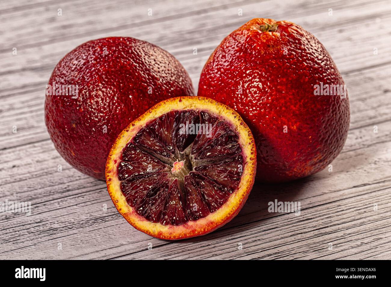 Tre arance di sangue esposte su tavole di legno intemperie, una tagliata a metà che rivela segmenti interni scuri color cremisi, vivaci fotografie agli agrumi Foto Stock