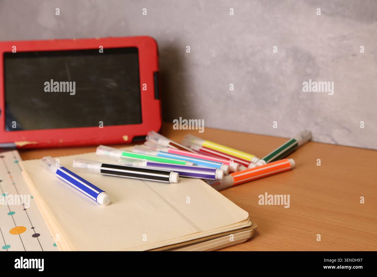 Una scrivania mostra una collezione di pennarelli di colori diversi sparsi intorno a un notebook chiuso. Un tablet si trova in una custodia rossa nelle vicinanze, creando un ambiente di lavoro informale. Foto Stock