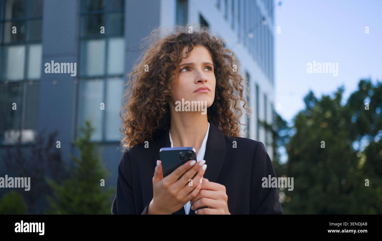 Perplessa donna d'affari caucasica che cerca un'applicazione gps per telefoni cellulari che cammina in città cerca di un luogo che guardi intorno all'edificio degli uffici Foto Stock