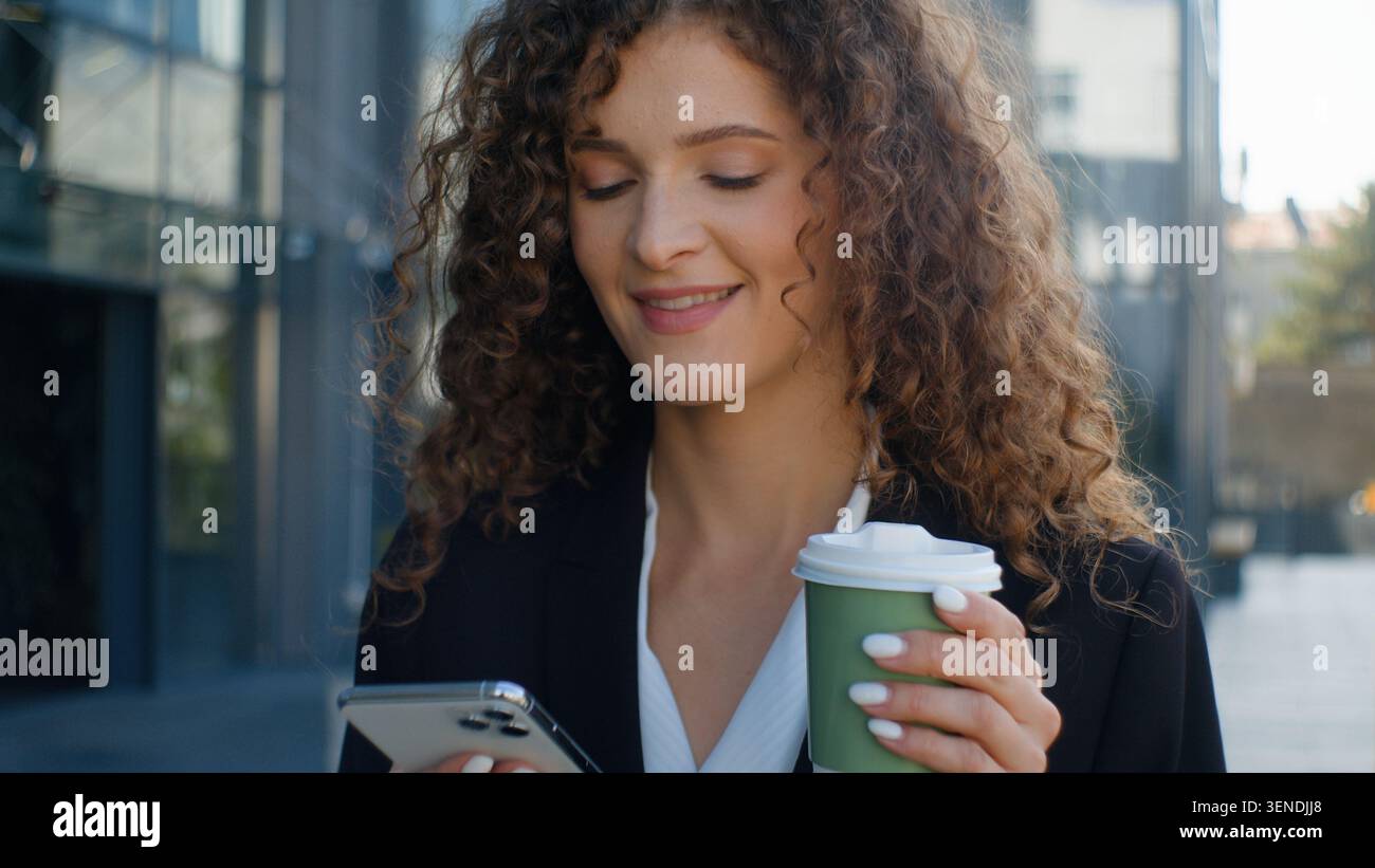 Gadget sorridente dipendente donna d'affari caucasica che cerca nel telefono cellulare bere caffè a piedi al lavoro al mattino in città donna d'affari ragazza femminile Foto Stock