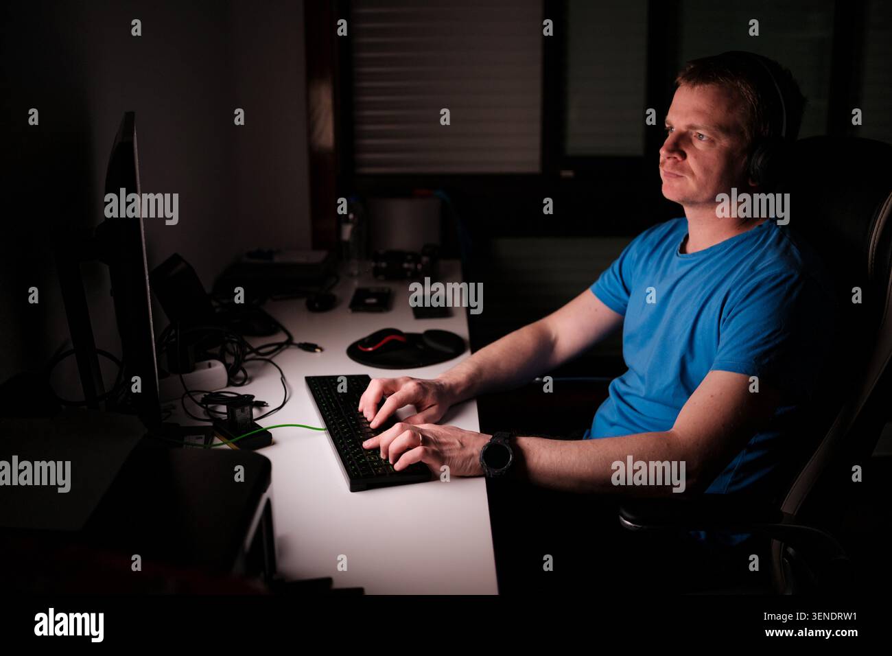 Uomo che indossa cuffie e t-shirt blu che scrive sulla tastiera in una stanza buia Foto Stock