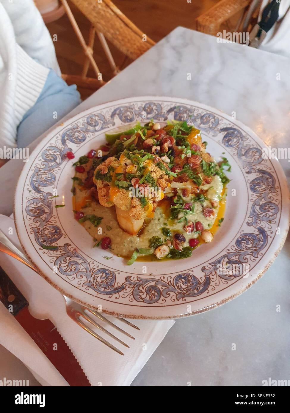 Bistecca di cavolfiore arrosto con salsa alle erbe verdi, noci tostate e semi di melograno su un piatto decorativo vintage, fotografata in un caffè in marmo Foto Stock