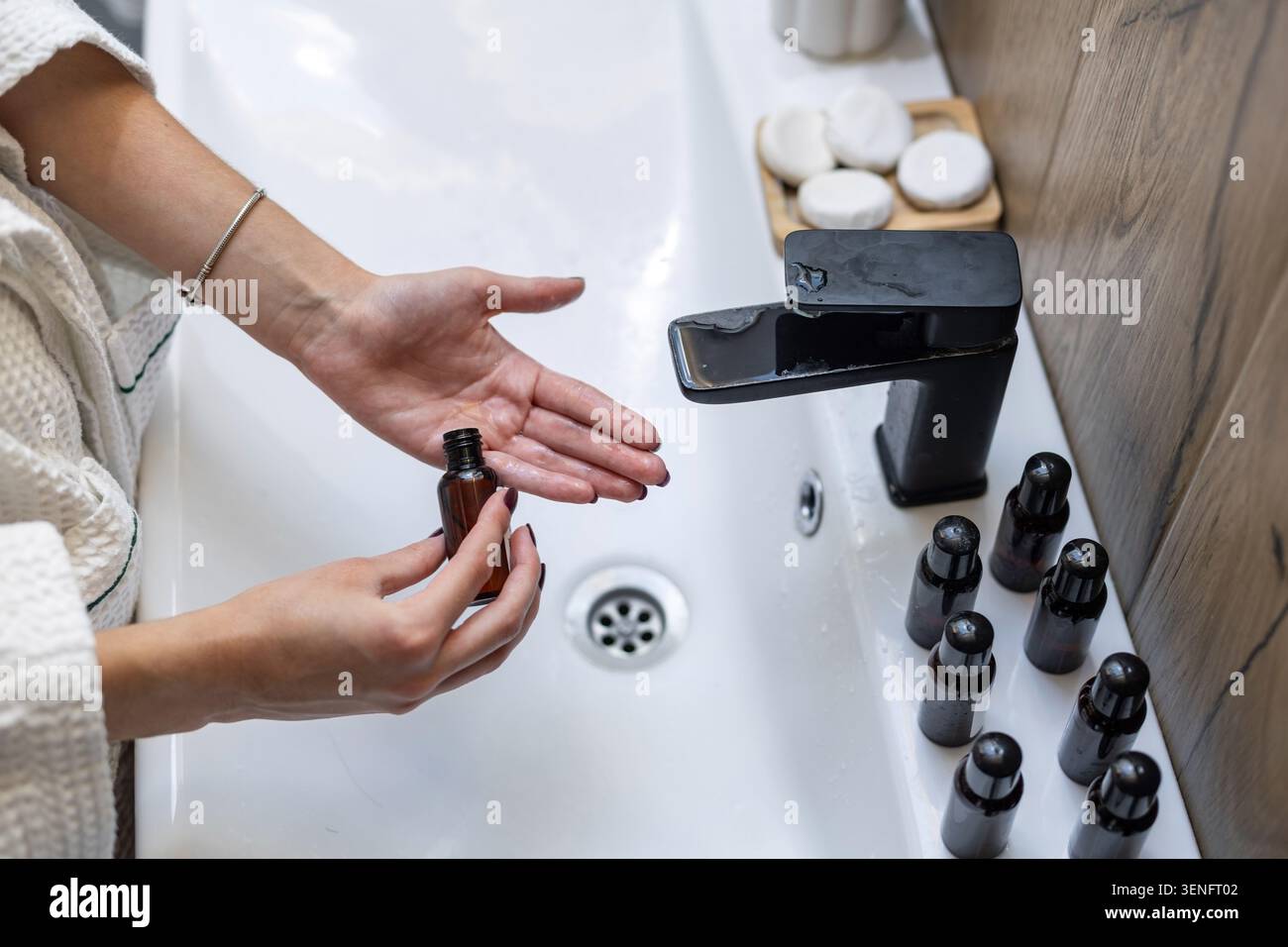 Donna che versa liquido per la cura della pelle nel palmo sopra il lavandino del bagno. Primo piano sopra la testa di una donna che tiene in mano una bottiglia di dermocosmesi ambrata aperta sopra il palmo bagnato Foto Stock