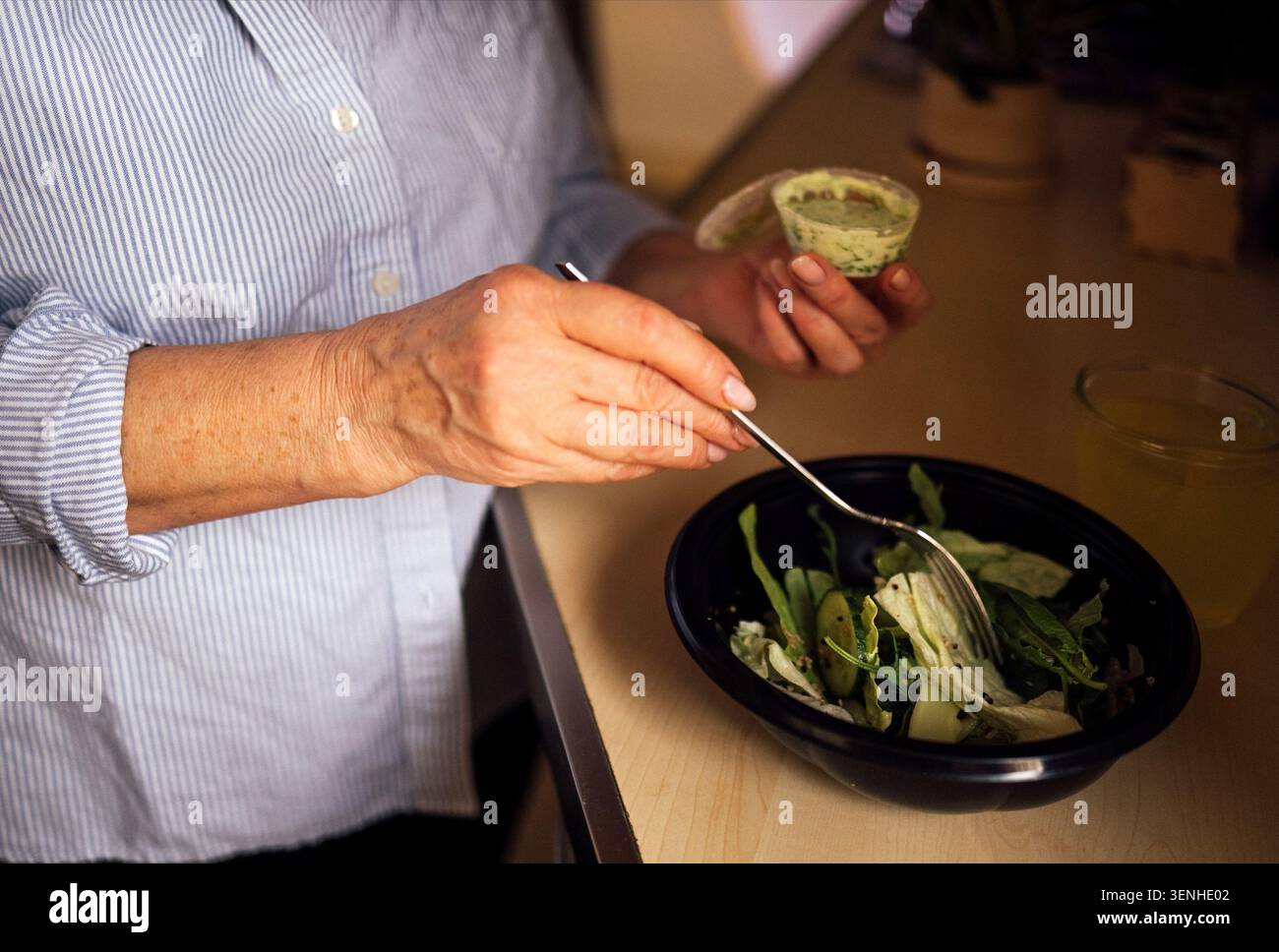 Una donna che indossa una camicia a righe blu mescola un'insalata fresca in una ciotola nera su un ripiano di legno, con una bevanda e un contenitore per condimenti visibili Foto Stock