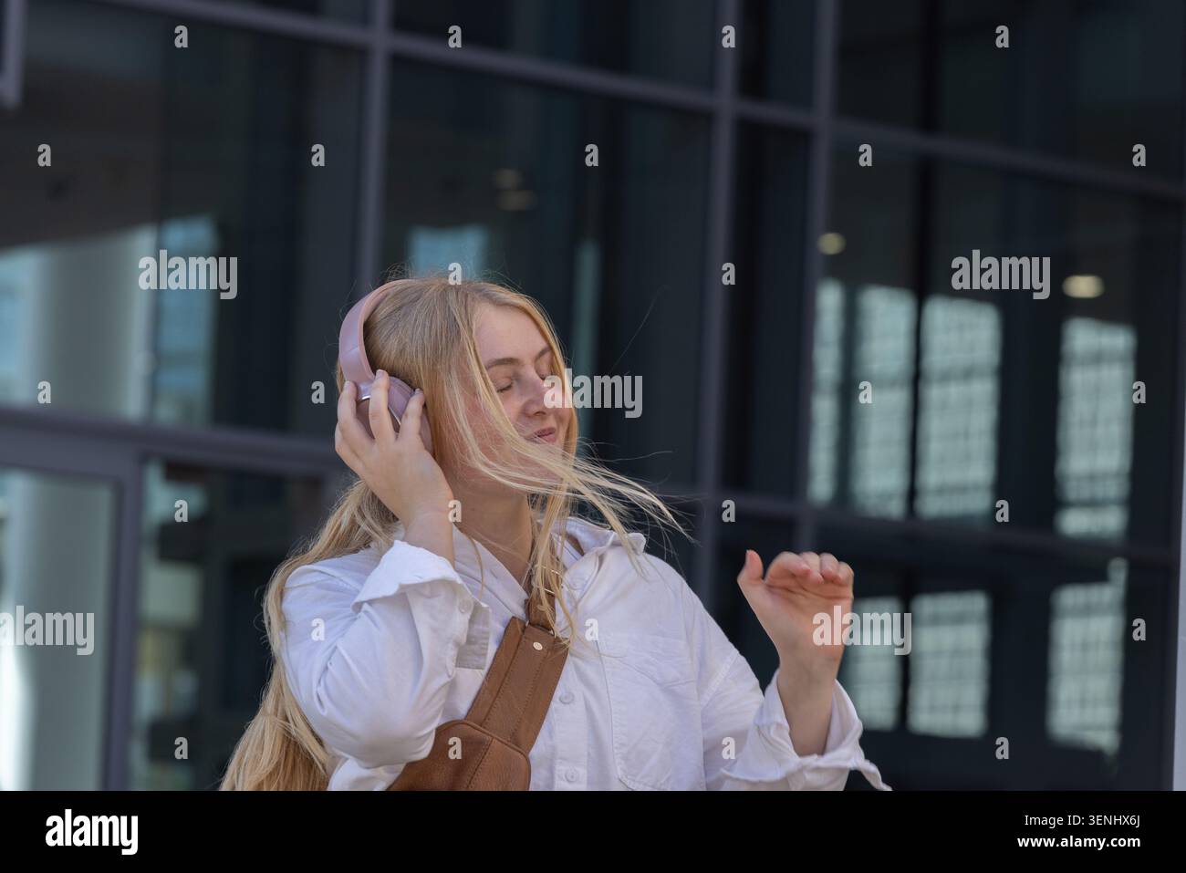 Donna in piedi di fronte a un edificio in vetro, che ascolta con le cuffie rosa e che tocca la tracolla della borsa Foto Stock