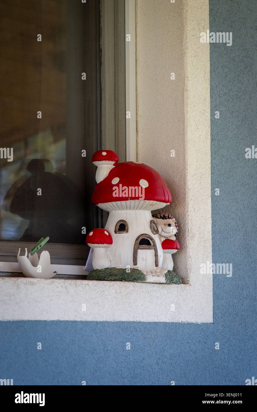 La statuetta decorativa rossa della casa dei funghi si trova su una finestra accanto a una parete esterna blu, aggiungendo un tocco stravagante del giardino a una tranquilla facciata domestica. Foto Stock