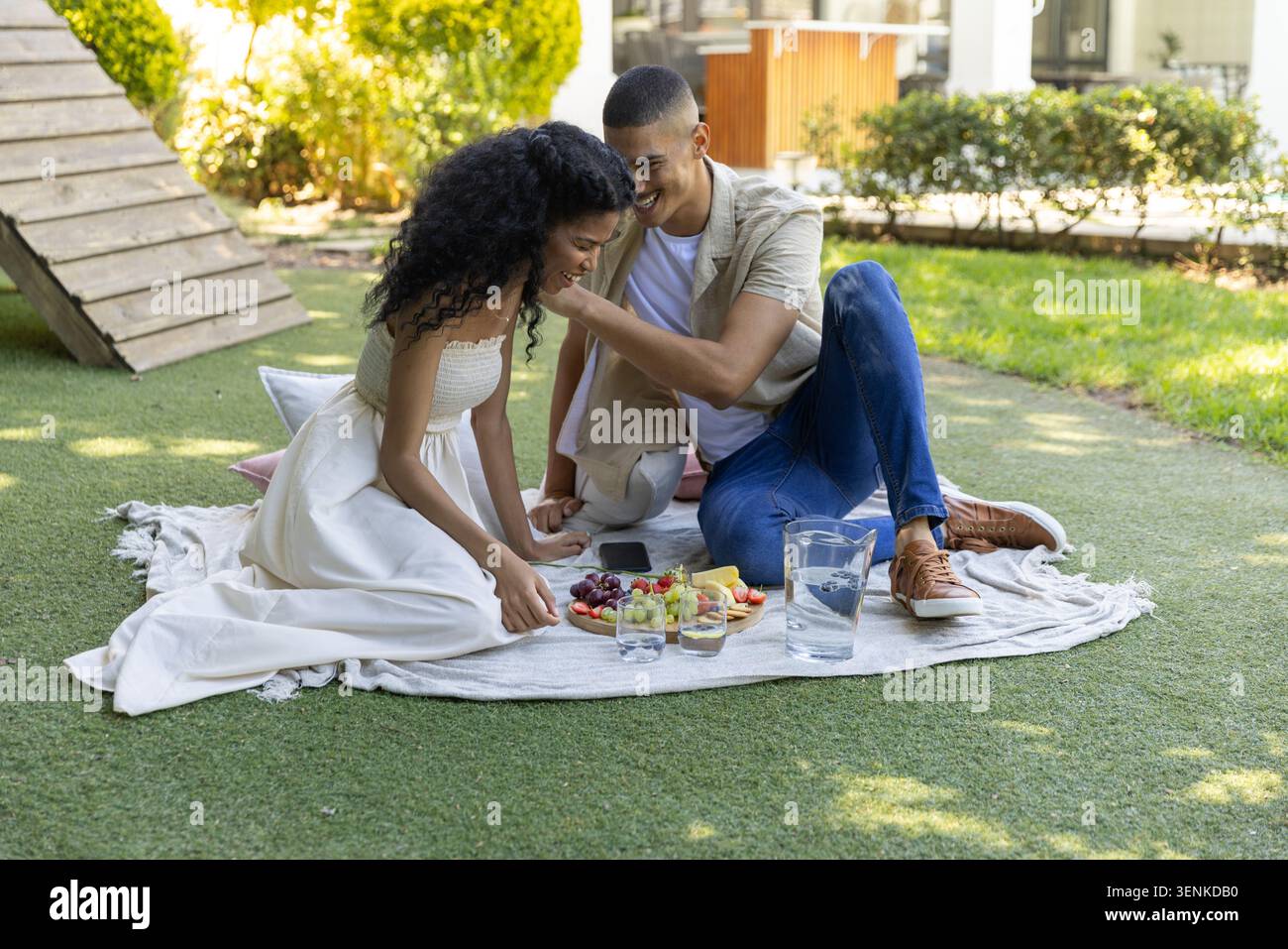 Coppia seduta su una coperta intrecciata nel cortile di casa condividendo un piatto di frutta con una caraffa, occhiali, smartphone Foto Stock