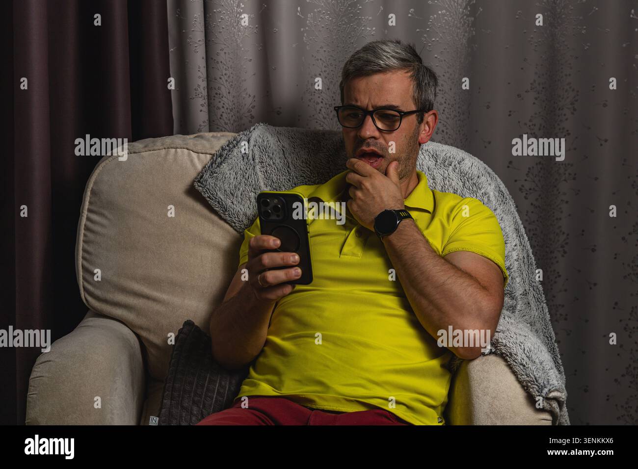 Un uomo seduto sul divano di casa, guardando il suo smartphone con un'espressione premurosa. Sembra stressato mentre legge messaggi e notifiche. Foto Stock