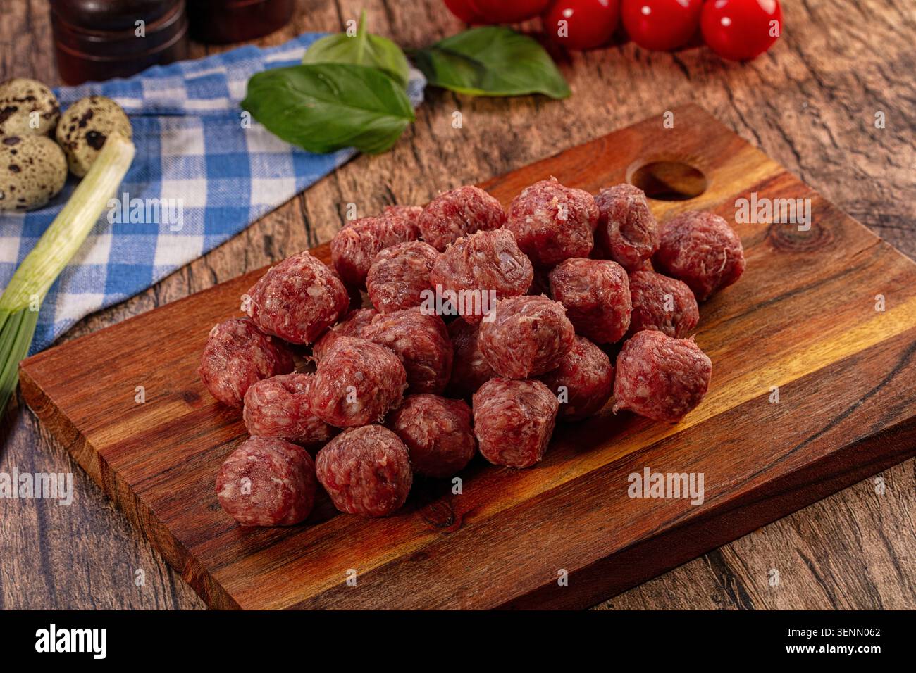 Un primo piano di polpette crude e crude disposte su un tagliere di legno scuro. Foglie di basilico verde fresco, pomodori rossi, uova di quaglia e un clo a scacchi Foto Stock