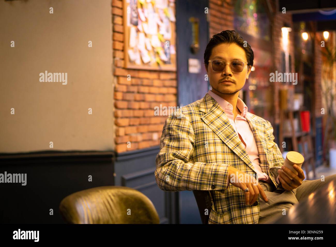 Uomo asiatico elegante in un blazer a quadri vintage e camicia rosa che si rilassa a un bancone di un bar di lusso e in un rigoglioso cortile giardino, ritratto sartoriale. Foto Stock