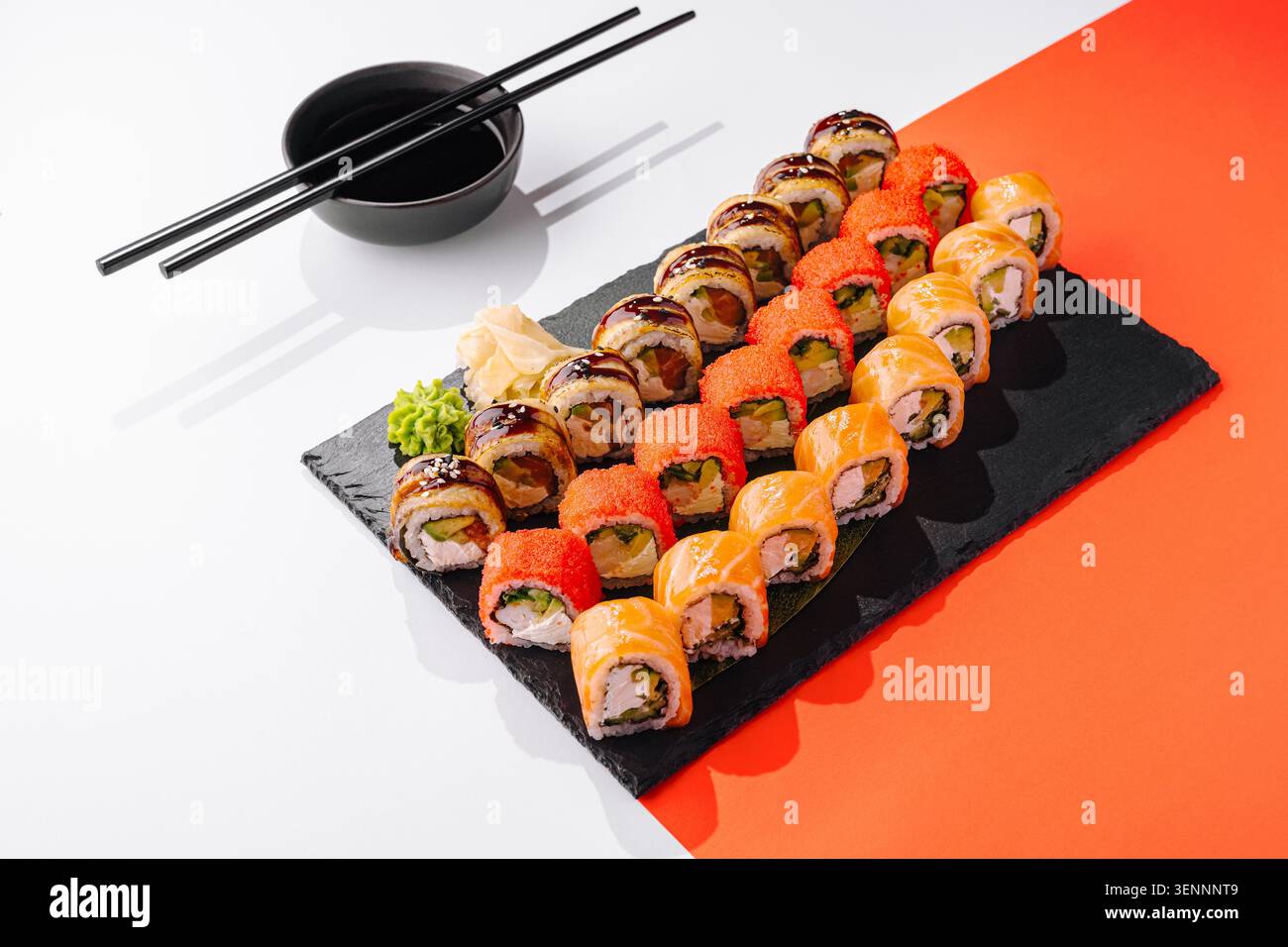 Gli involtini di sushi disposti ordinatamente su un piatto nero sono pronti da mangiare. Due tipi di rotoli sono dotati di guarnizioni colorate. Le bacchette e una piccola ciotola di salsa di soia si trovano nelle vicinanze. Foto Stock