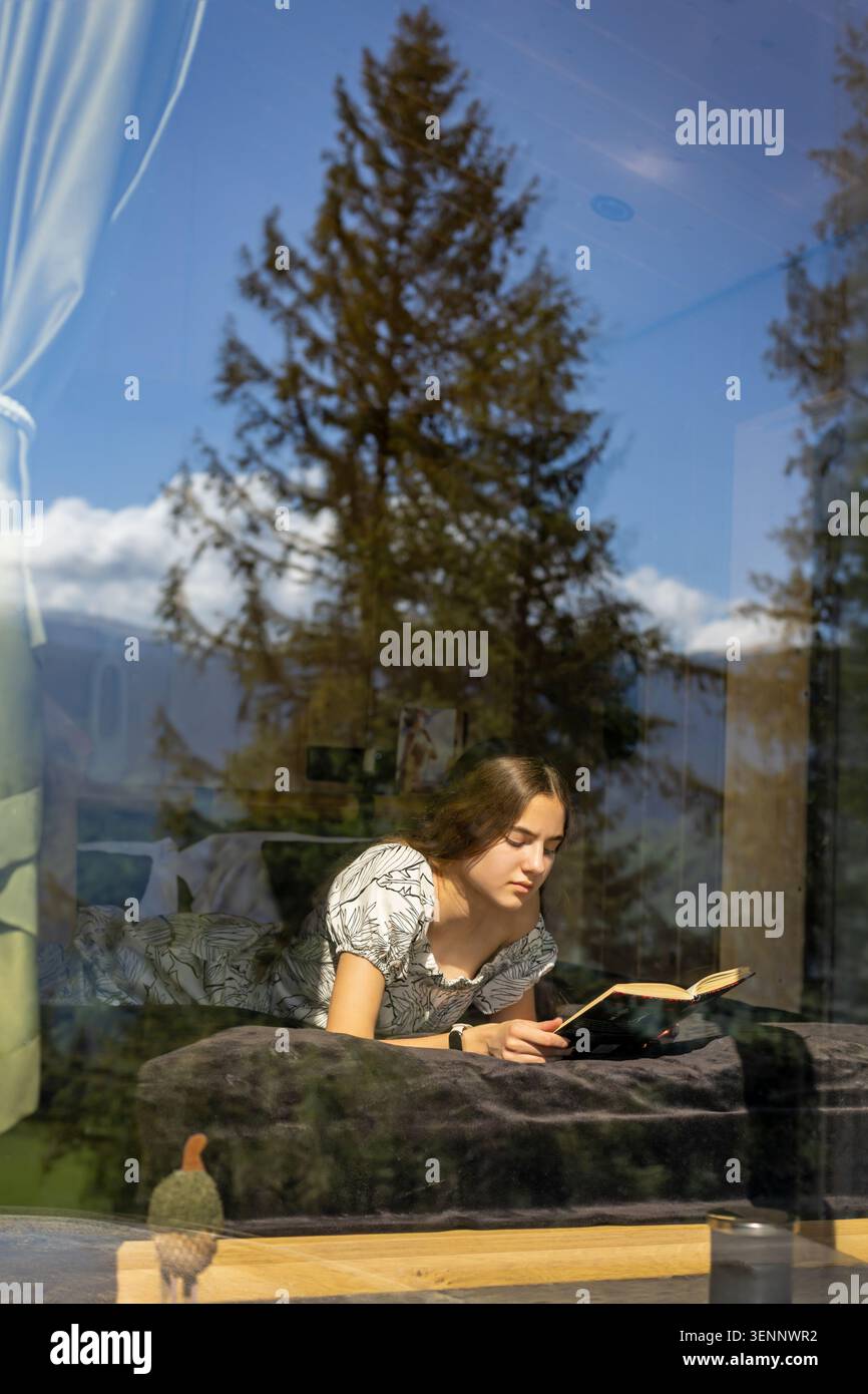 Donna che legge in cabina dietro la finestra della foresta riflettente. Una giovane donna giace su un letto scuro leggendo un libro aperto all'interno di una cabina di legno, vista attraverso Foto Stock