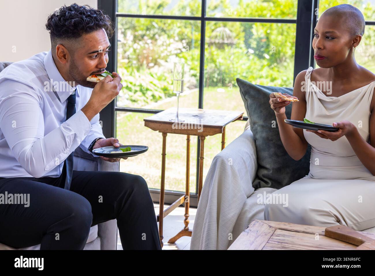 Coppia afroamericana seduta a casa in abito formale mangiando panini a faccia aperta su piatti neri Foto Stock