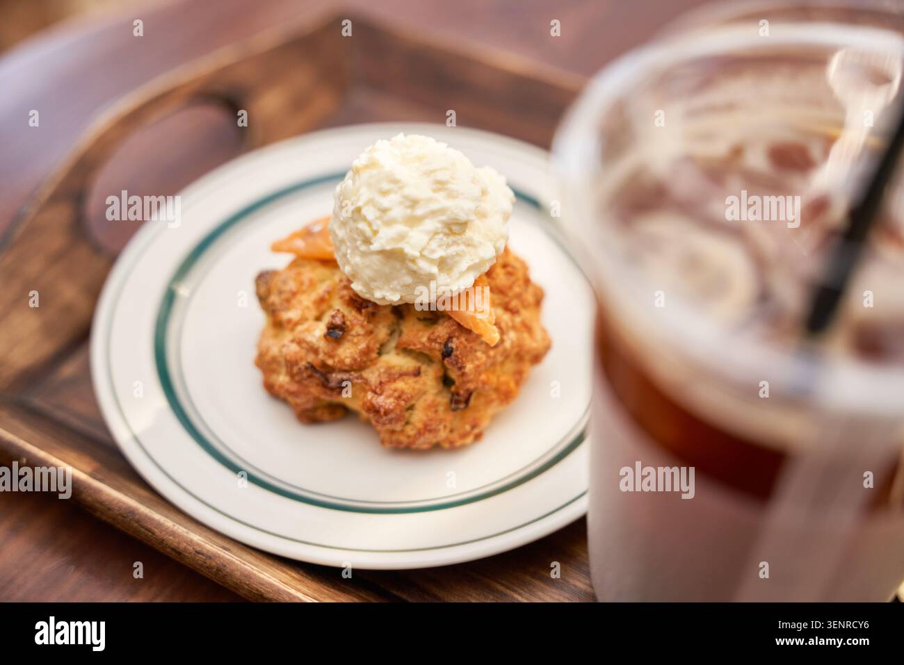 Una pasticceria al forno con una ricca panna si trova su un vassoio di legno vicino a una bevanda ghiacciata sfocata, catturando perfettamente un intimo e rilassante momento da dessert Foto Stock