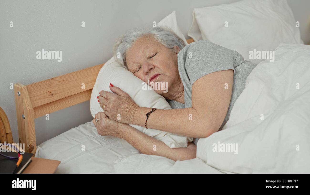Una donna anziana abbraccia un cuscino mentre dorme su un letto in una camera da letto, distesa sul fianco con gli occhi chiusi e le mani che stringono il cuscino; comfort serenità. Foto Stock