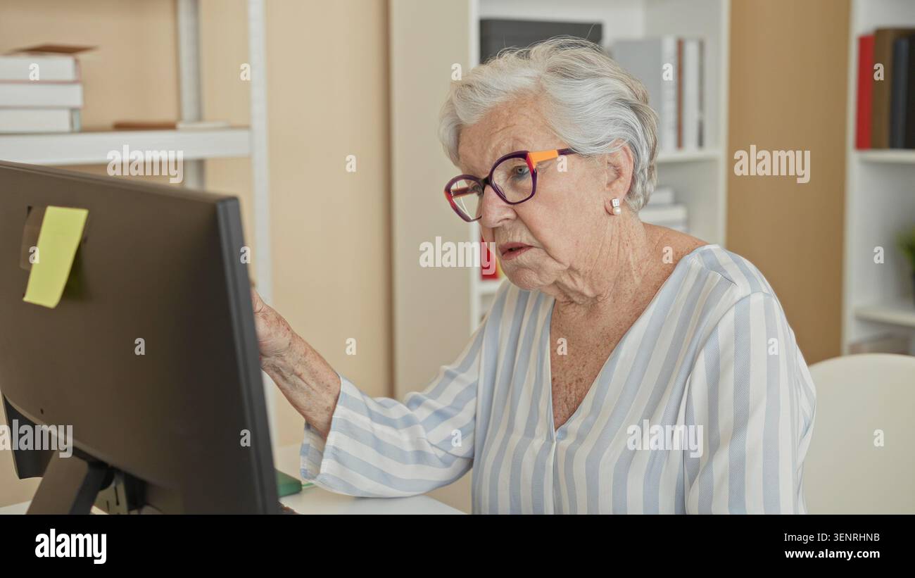 La donna anziana punta il dito indice sul monitor del computer, la mano è visibile, porta gli occhiali in un ufficio edile; indipendenza dalla concentrazione. Foto Stock