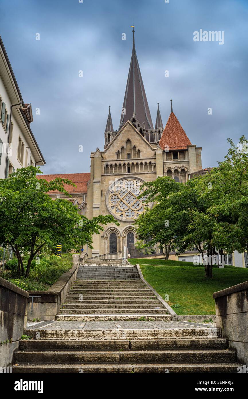 I gradini in pietra conducono a una chiesa storica con alte guglie, una grande rosetta e alberi verdi sotto un cielo nuvoloso in un tranquillo ambiente cittadino europeo. Foto Stock