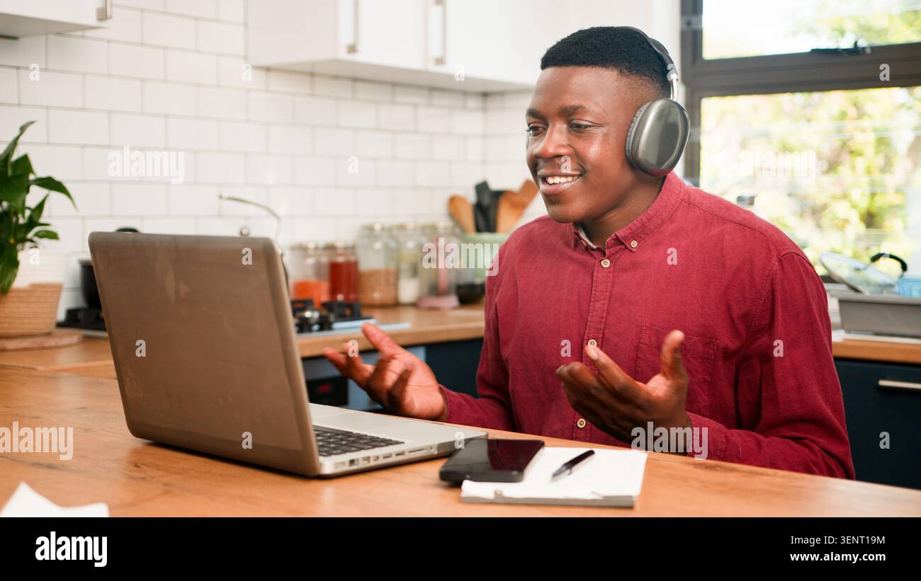 Riunione aziendale remota con un uomo sorridente che indossa le cuffie alla scrivania della cucina di casa per la consulenza finanziaria Foto Stock