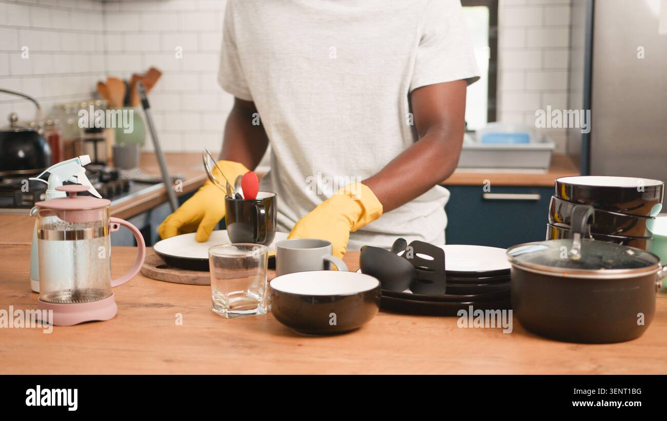 Persona che lava i piatti in cucina indossando guanti gialli con pentole e tazze impilate Foto Stock
