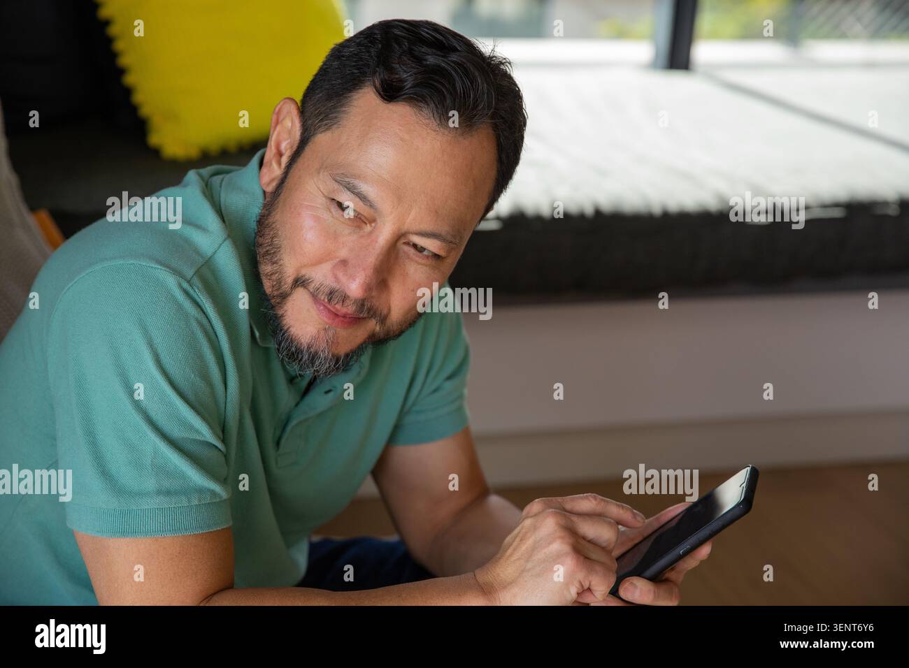 Uomo asiatico maturo seduto su panchina bassa vicino alla finestra, toccando telefono nero, polo verde acqua, cuscino giallo Foto Stock