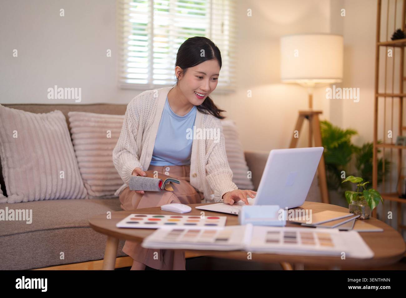 Giovane interior designer asiatico che lavora su progetti di design di mobili con campioni di materiale e computer nel soggiorno Foto Stock