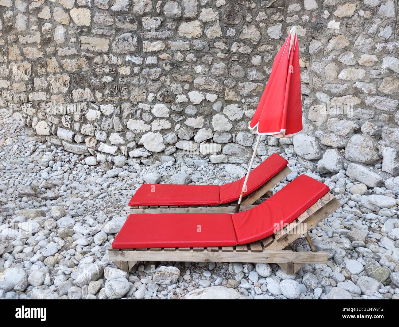 Vecchie sdraio rosse e ombrellone su una spiaggia di ciottoli contro un muro di pietra. Splendida vacanza per rilassarsi all'ombra in riva al mare. Copia sp Foto Stock
