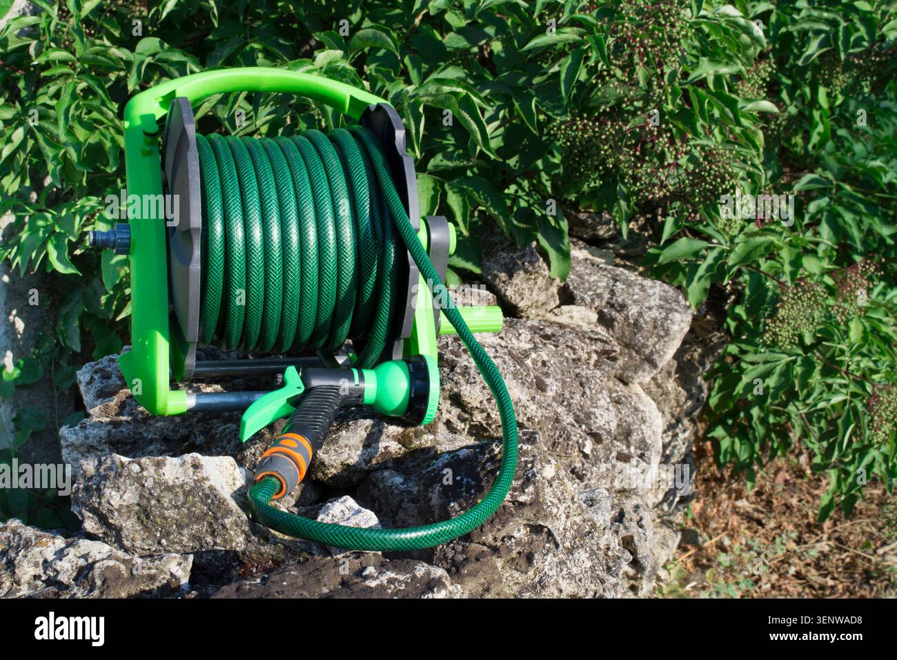Una bobina con un tubo per innaffiare verde con una pistola ad acqua si appoggia su pietre sullo sfondo di piante verdi. Casa e giardinaggio. Foto Stock