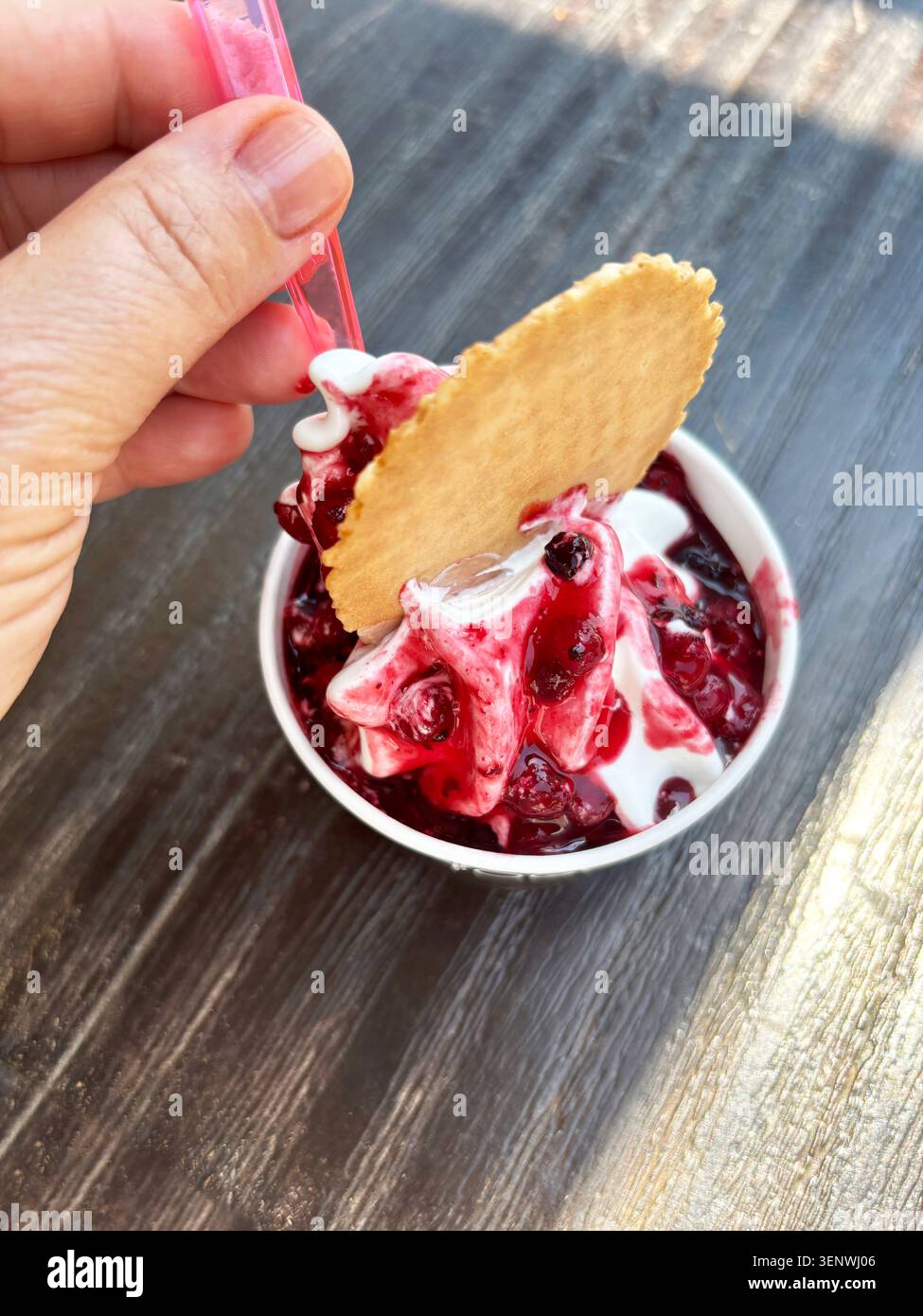 Tazza di yogurt surgelato servito con salsa dolce mista di frutti di bosco e un cialda con cono di waffle, tenuto da un cliente all'aperto su un tavolo di legno. Foto Stock