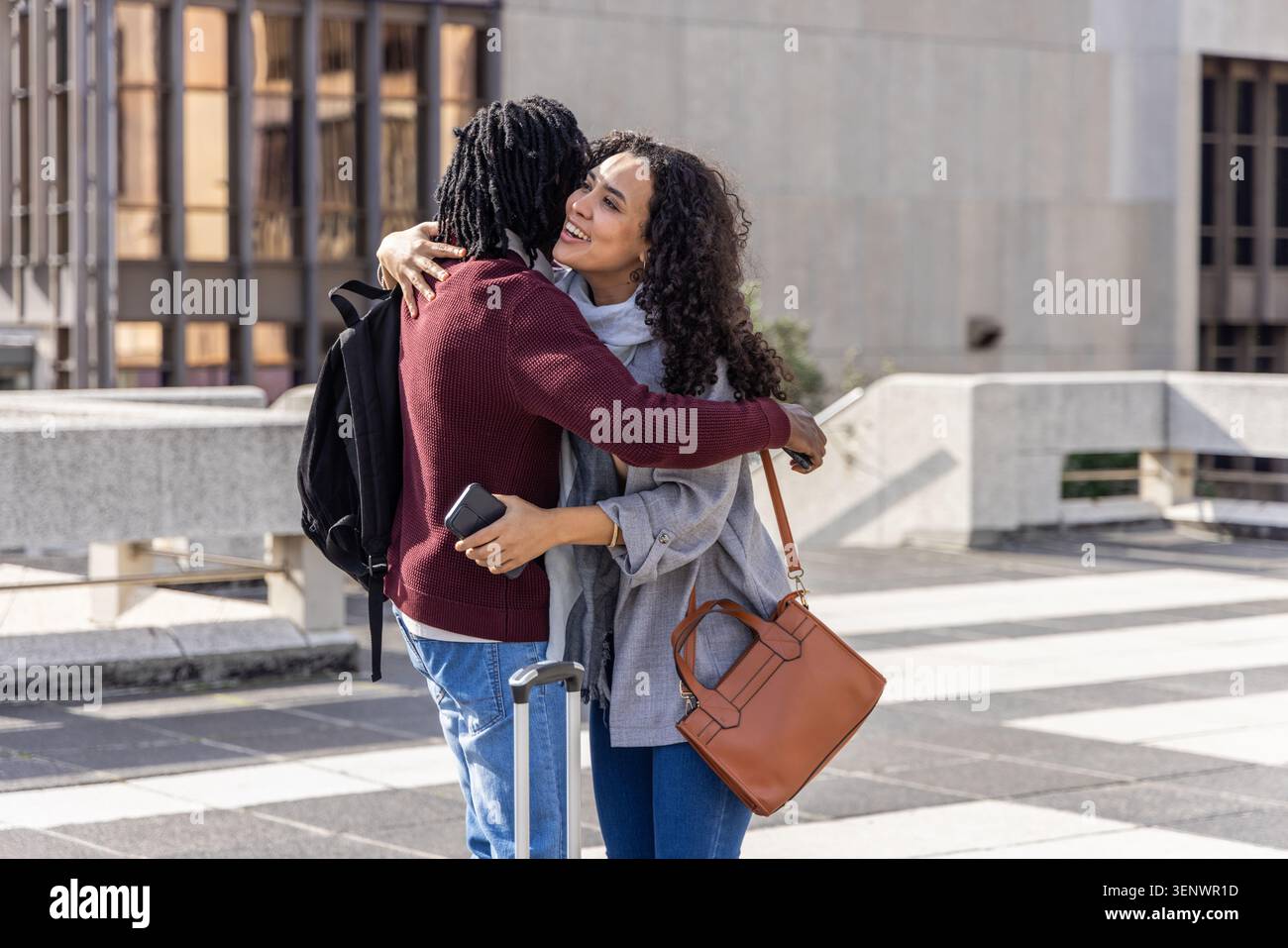 Abbinalo a maglia Bordeaux e cappotto grigio che abbracciano Urban plaza, tenendo in mano telefoni e zaino Foto Stock