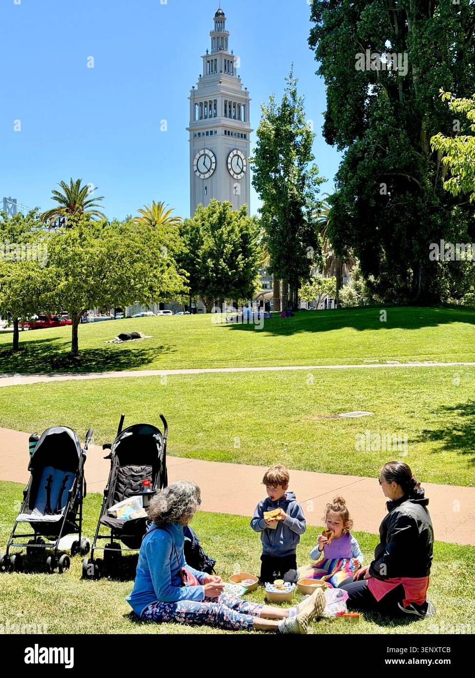 Nonne e nipoti che fanno un picnic in un parco a San Francisco, California. Torre dell'orologio di 245 metri circa in cima allo storico Ferry Building sullo sfondo. - Immagine stock catturata con smartphone