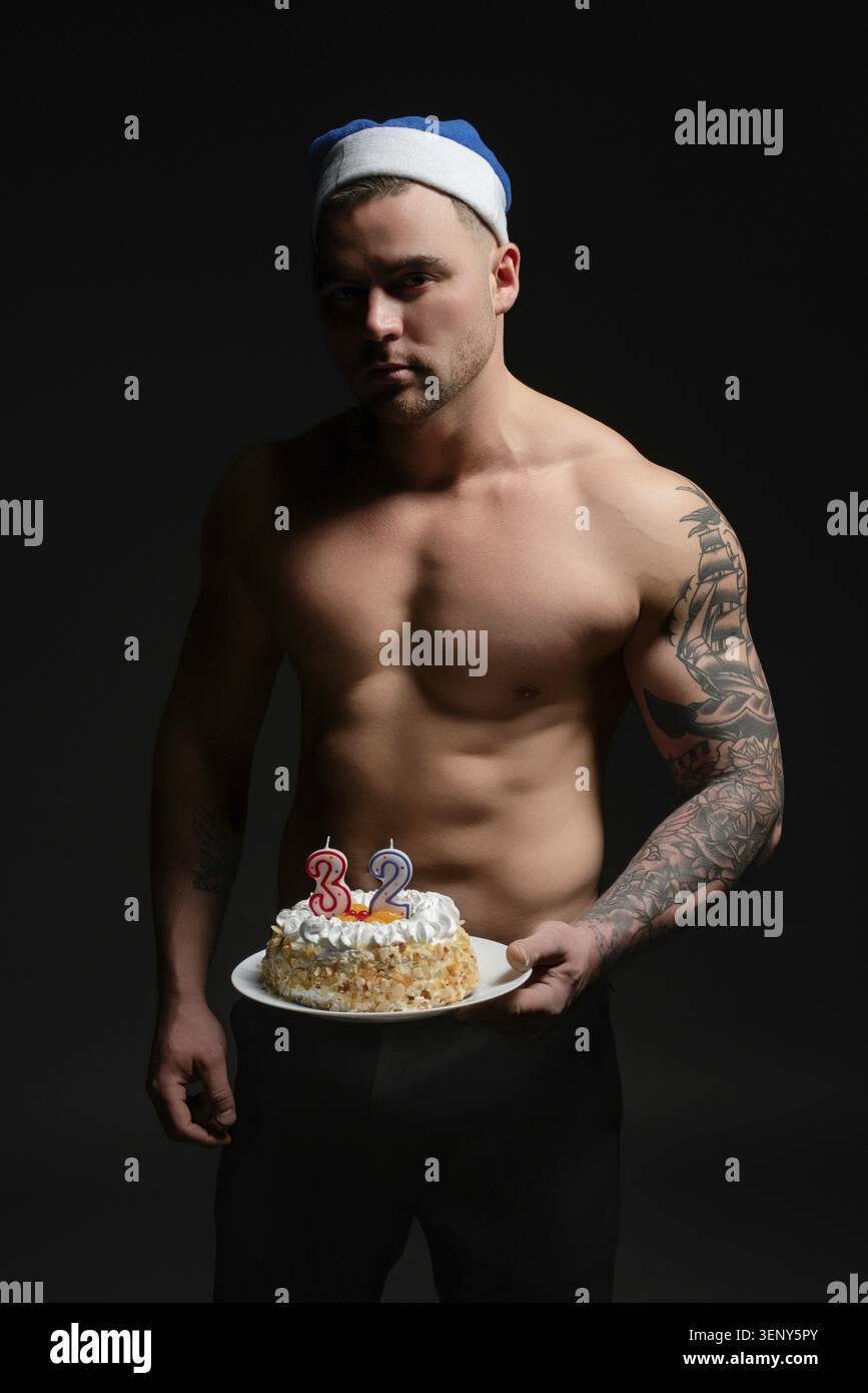 L'uomo con i tatuaggi si trova in una luce soffusa e tiene una torta di compleanno, festeggiando con orgoglio i 32 anni di età Foto Stock