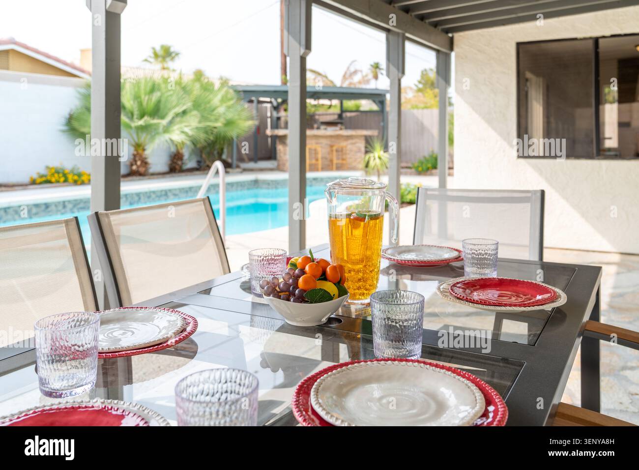 Set da pranzo all'aperto con tavolo in vetro, sedie a rete, piatti, bicchieri e una caraffa di tè freddo con sfondo piscina Foto Stock