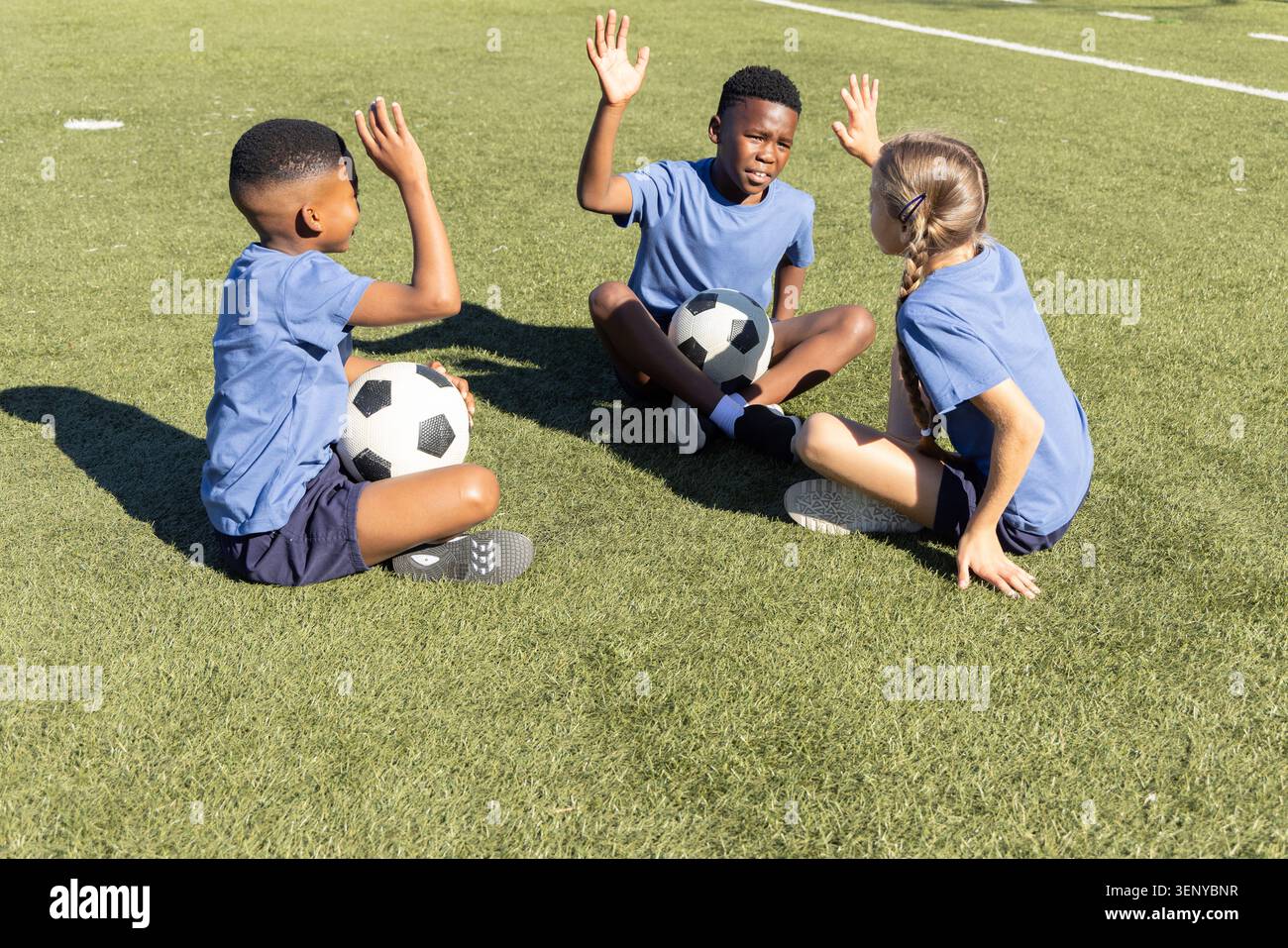 Bambini diversi seduti su erba erbosa con magliette blu che tengono le palle da calcio alzando le mani sotto il sole Foto Stock