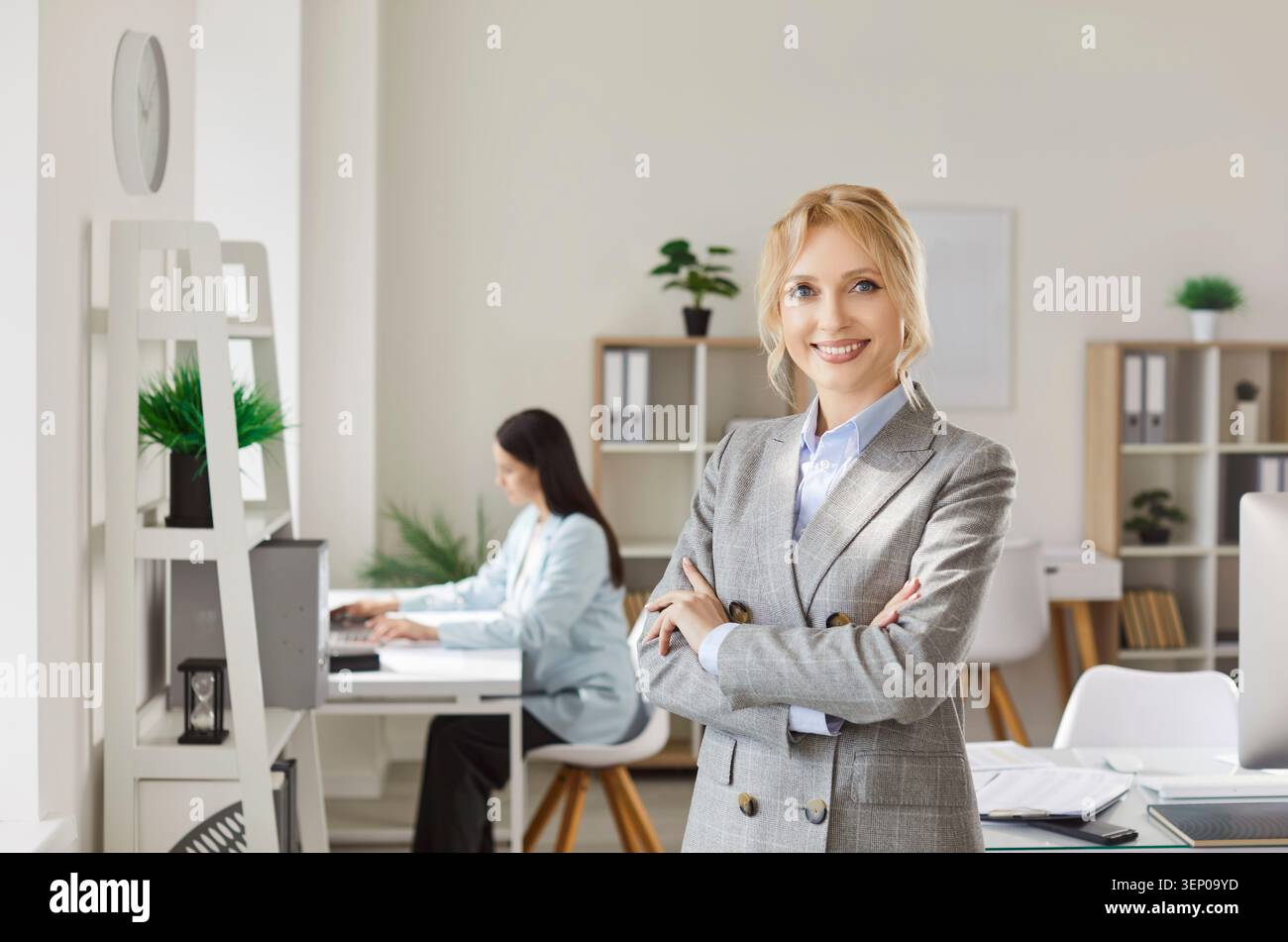 Ritratto di una donna d'affari bionda sorridente e sicura in tuta grigia in piedi con le braccia piegate in ufficio. Foto Stock