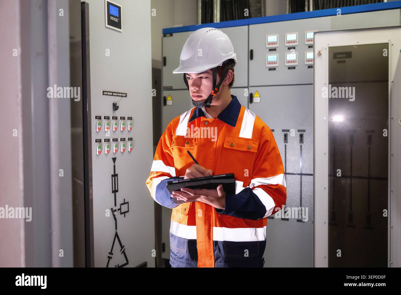 Giovane ingegnere industriale asiatico in gilet di sicurezza arancione e supporti per elmetto bianco. contiene tablet e penna. esamina lo schermo con attenzione. Pannelli elettrici lin Foto Stock